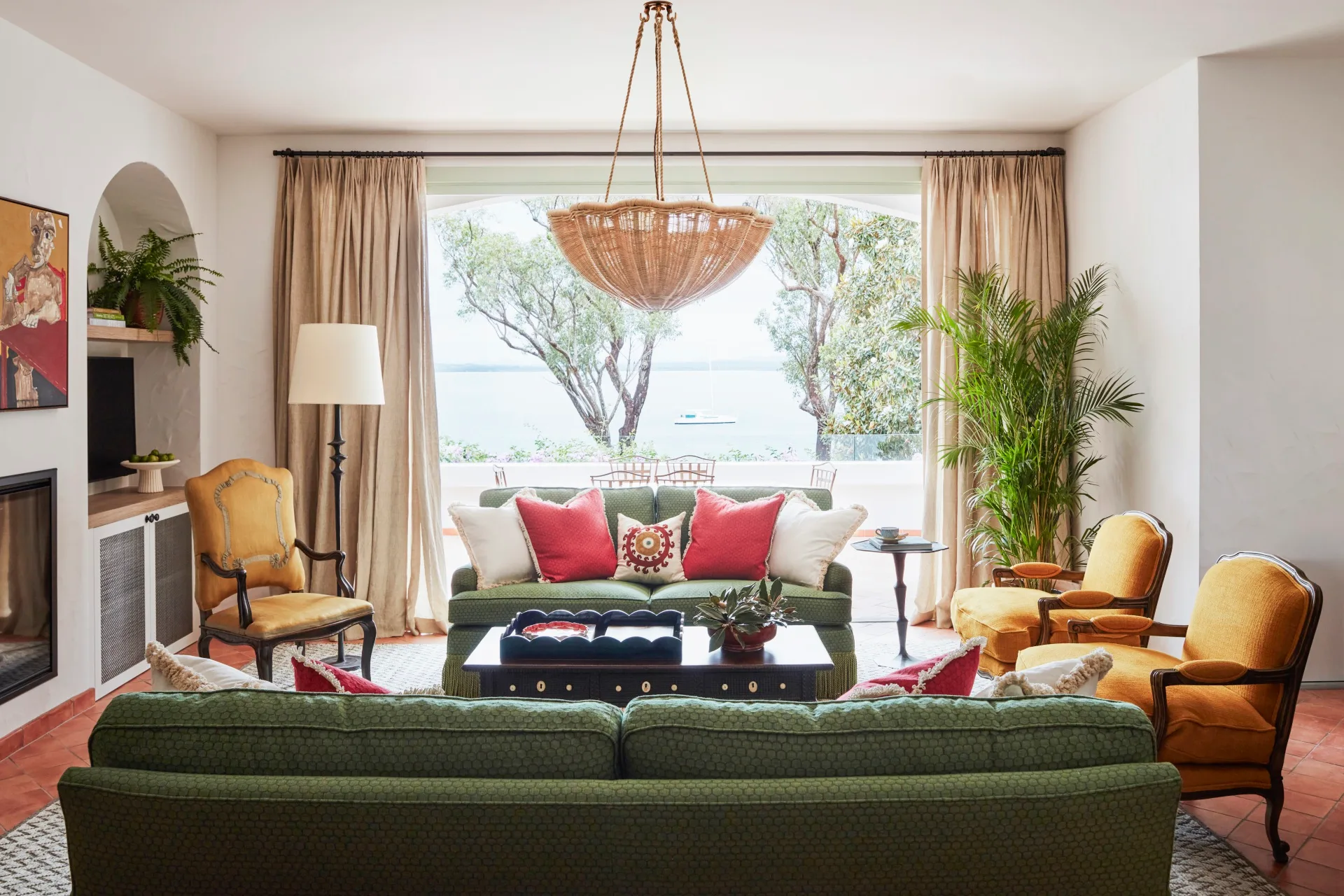 Elegant living room with green couches, red pillows, large window view of trees and water, wicker chandelier, and plants.
