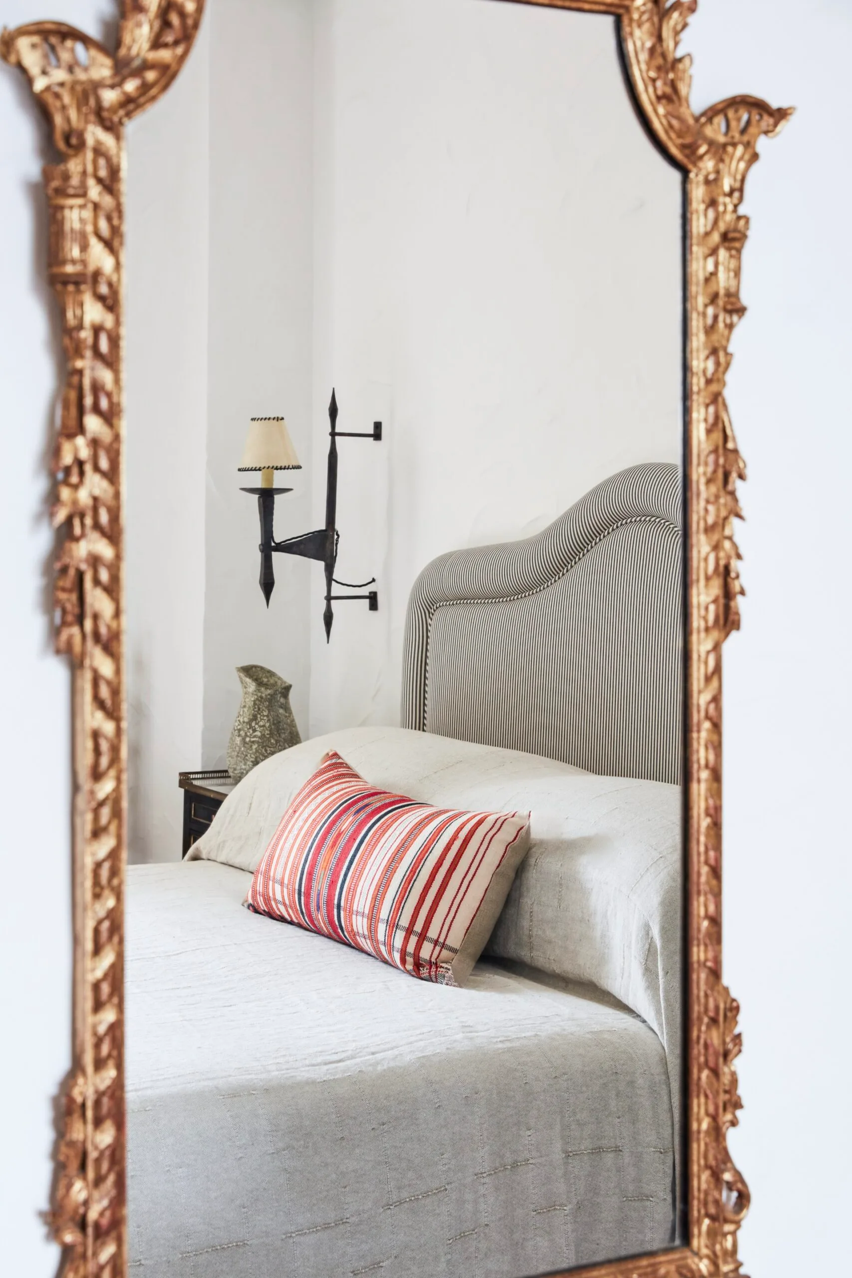Reflection of a bed with a striped headboard and red-striped pillow in an ornate gold-framed mirror.