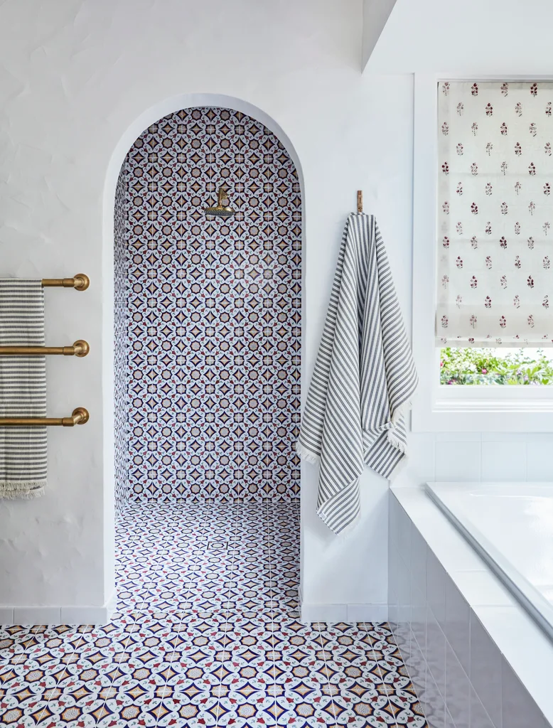 Bathroom with patterned tiles, archway shower, striped towels, brass hardware, and floral patterned blinds. The floor tiles carry up the wall in the shower area.