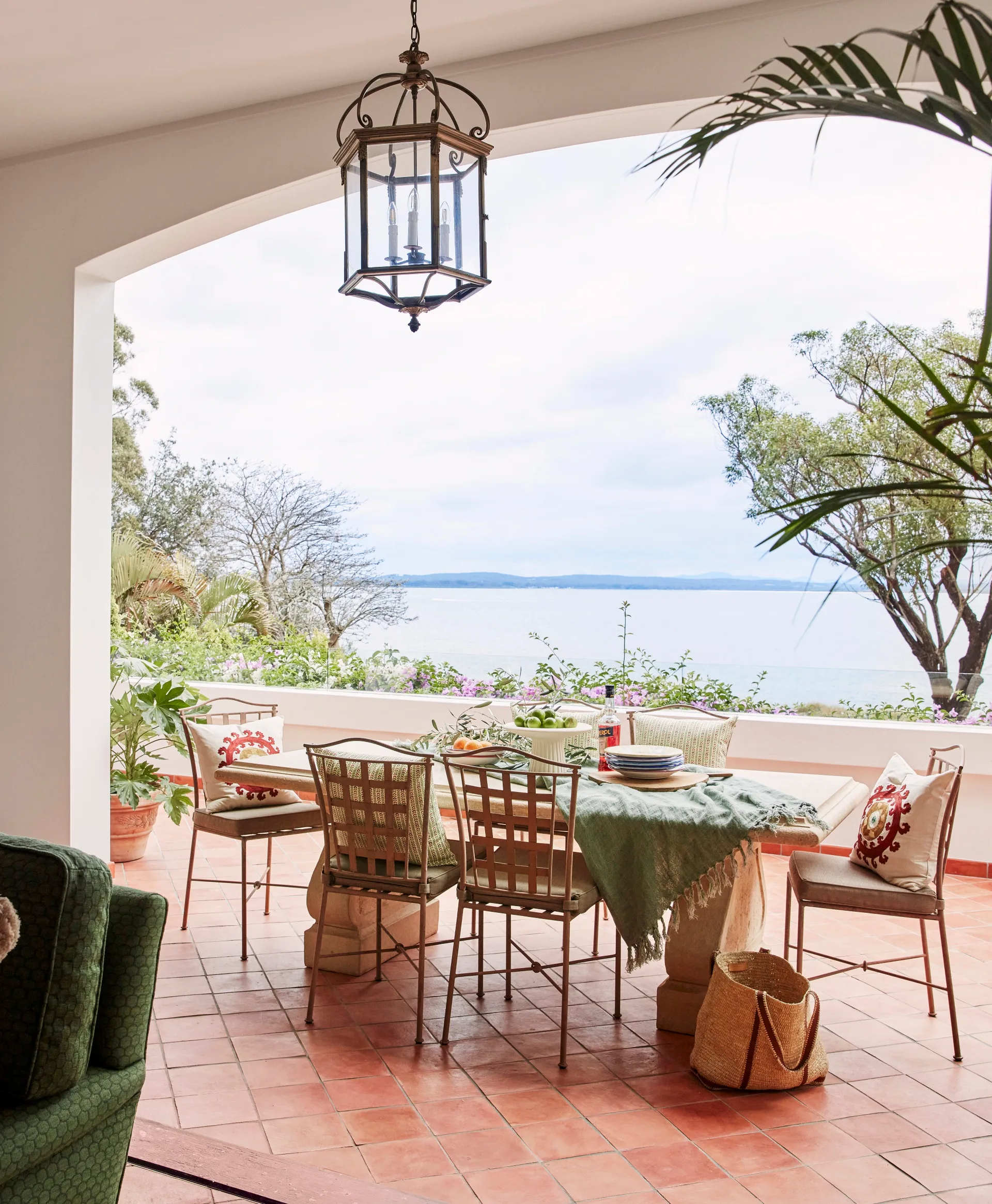 Covered patio with dining set, green throw blanket, and views of trees and a calm lake in the background. The terrace has a Mediterranean style with terracotta floor tiles.