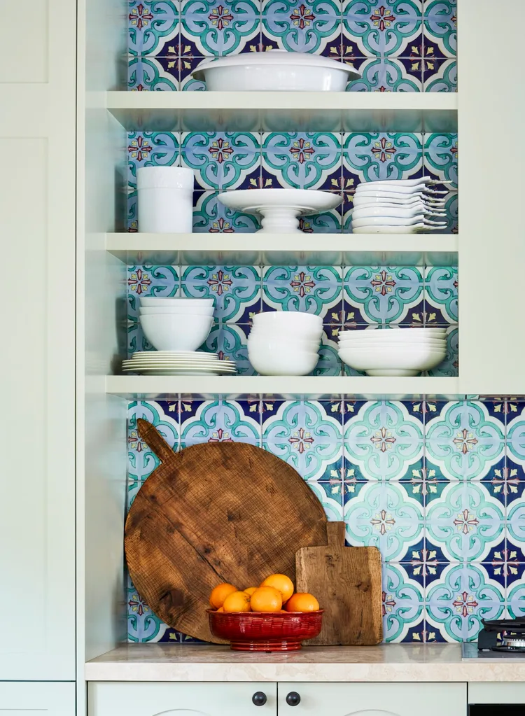 Open kitchen shelves with colorful tile backsplash, white dishes, wooden cutting boards, and a bowl of oranges. The cabinetry is green. There is a Mediterranean and European style.