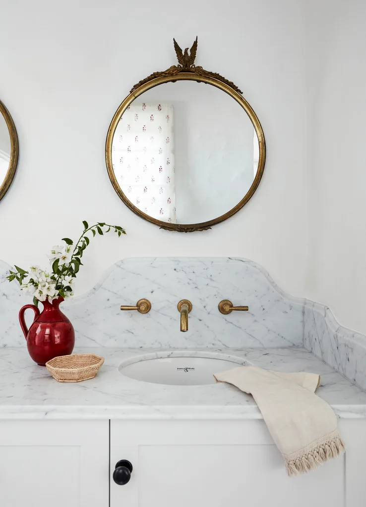Elegant bathroom sink with marble countertop, gold fixtures, ornate mirror, red vase with flowers, and woven basket.