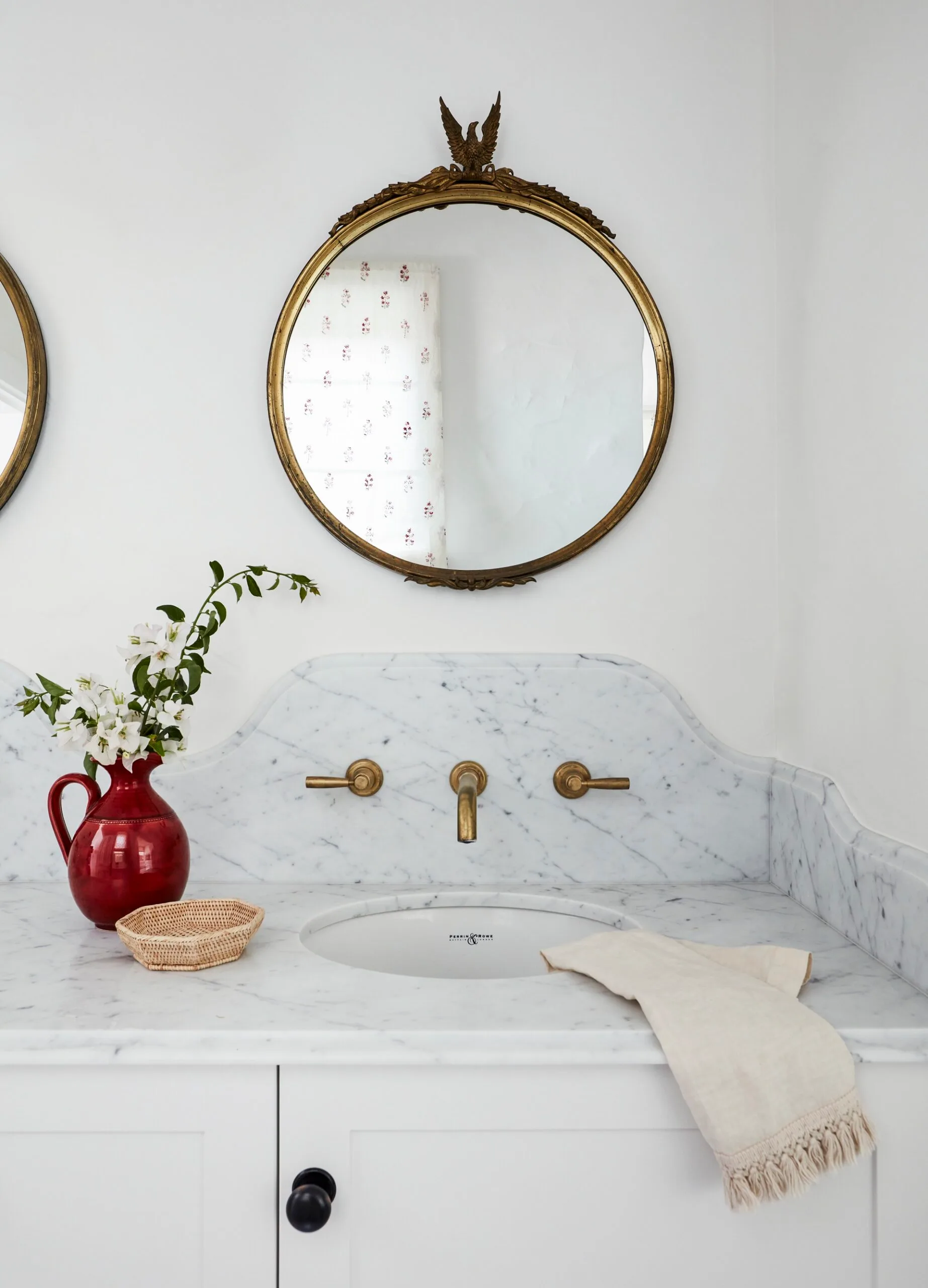 Elegant bathroom sink with marble countertop, gold fixtures, ornate mirror, red vase with flowers, and woven basket.