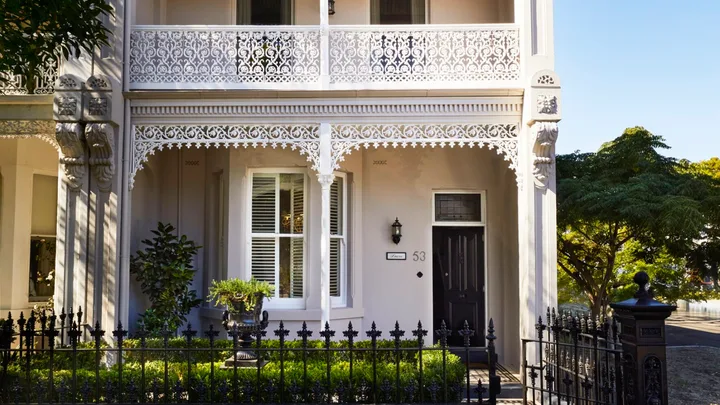 Elegant two-story Victorian terrace house with ornate white iron lacework and lush garden, under a clear blue sky.