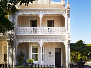 Elegant two-story Victorian terrace house with ornate white iron lacework and lush garden, under a clear blue sky.