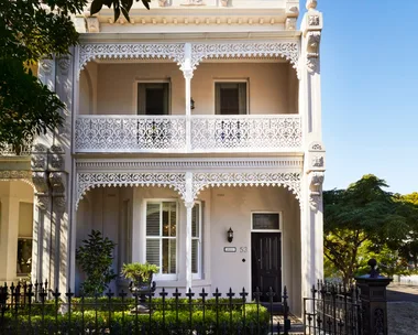 Elegant two-story Victorian terrace house with ornate white iron lacework and lush garden, under a clear blue sky.