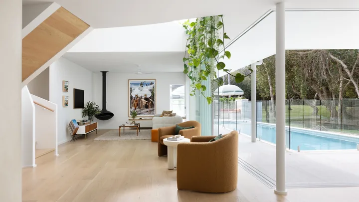 Modern living room with beige chairs, large windows, greenery, and a view of a pool and garden outside.