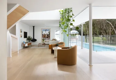 Modern living room with beige chairs, large windows, greenery, and a view of a pool and garden outside.