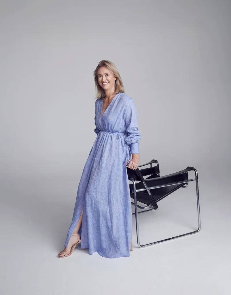an Australian designer in a blue dress leans on a modern chair, standing in a minimalist studio setting.