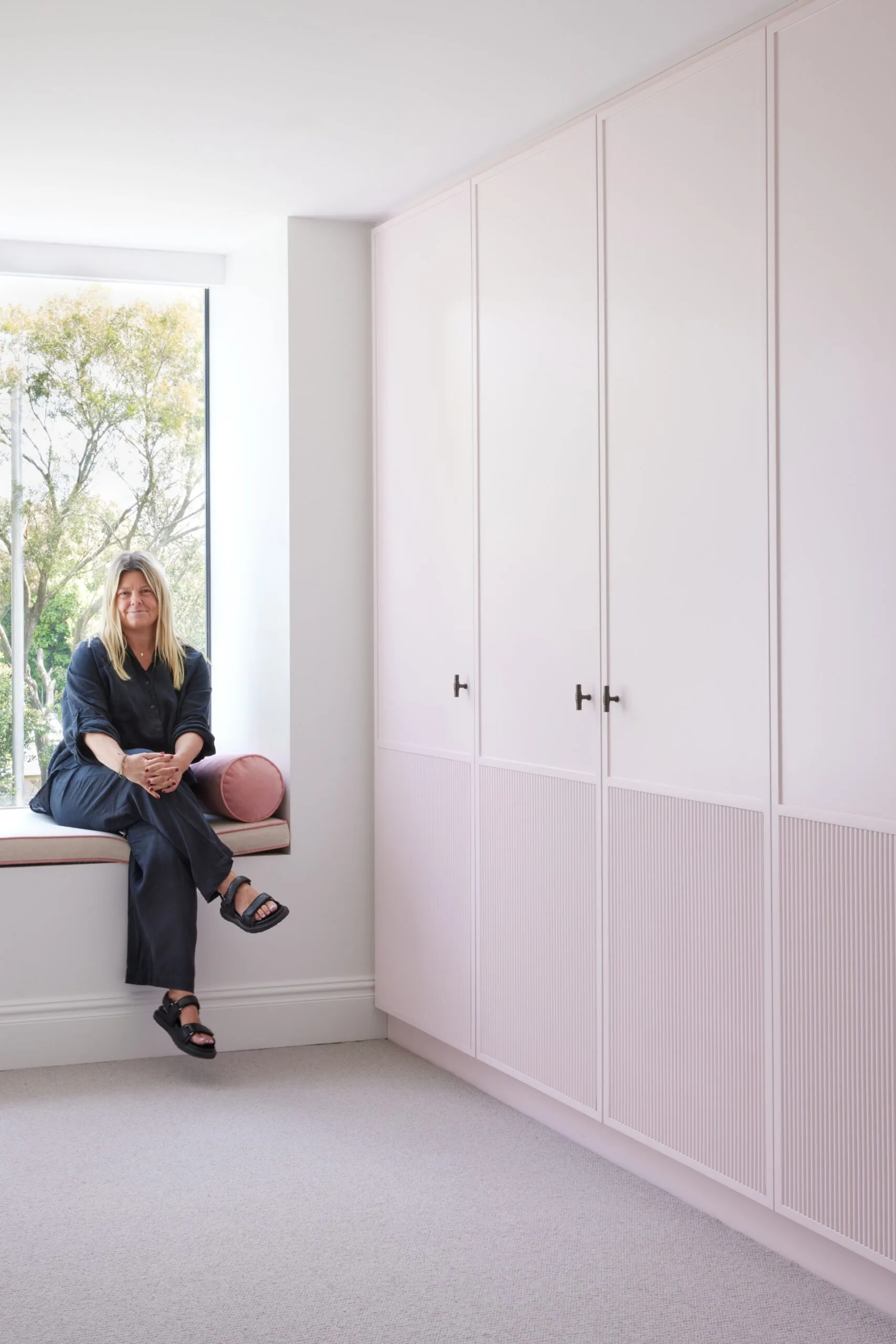 an Australian designer in dark clothing sits on a window bench beside pale pink cabinets, with trees visible outside.