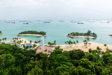 Aerial view of Sentosa Beach, Singapore, with palm trees and ships in the background on a sunny day.