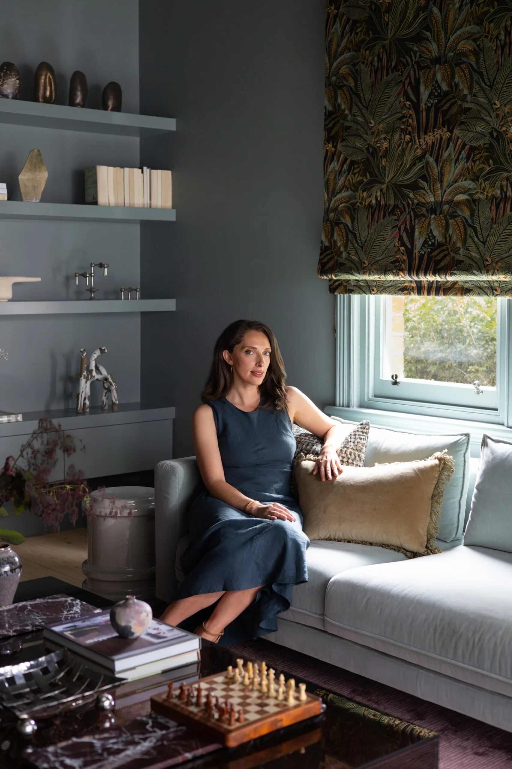 an Australian designer in blue dress sits on sofa in elegant living room with shelves, chessboard, and patterned curtains.