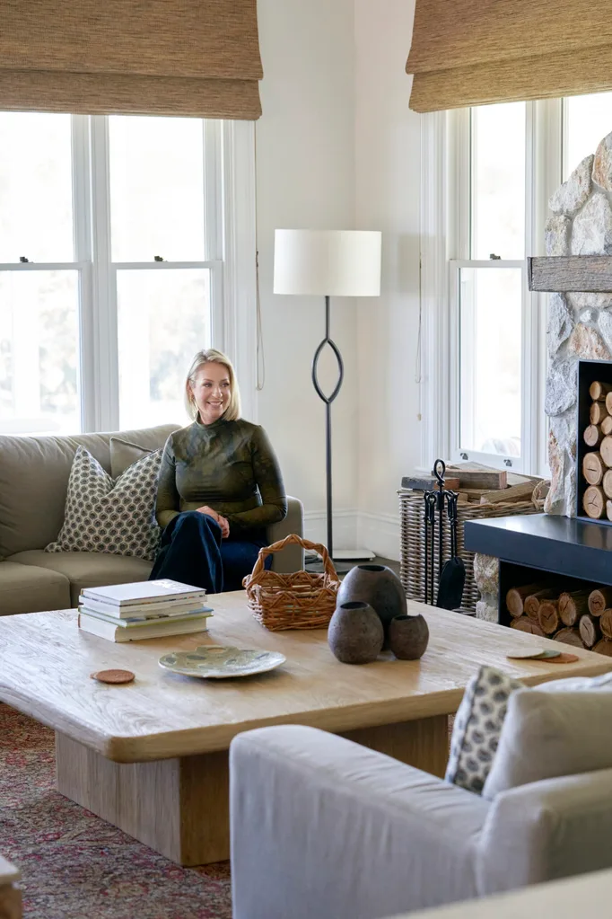 an Australian designer sitting on sofa in cozy living room with wooden table, lamps, and fireplace; books and basket on table.