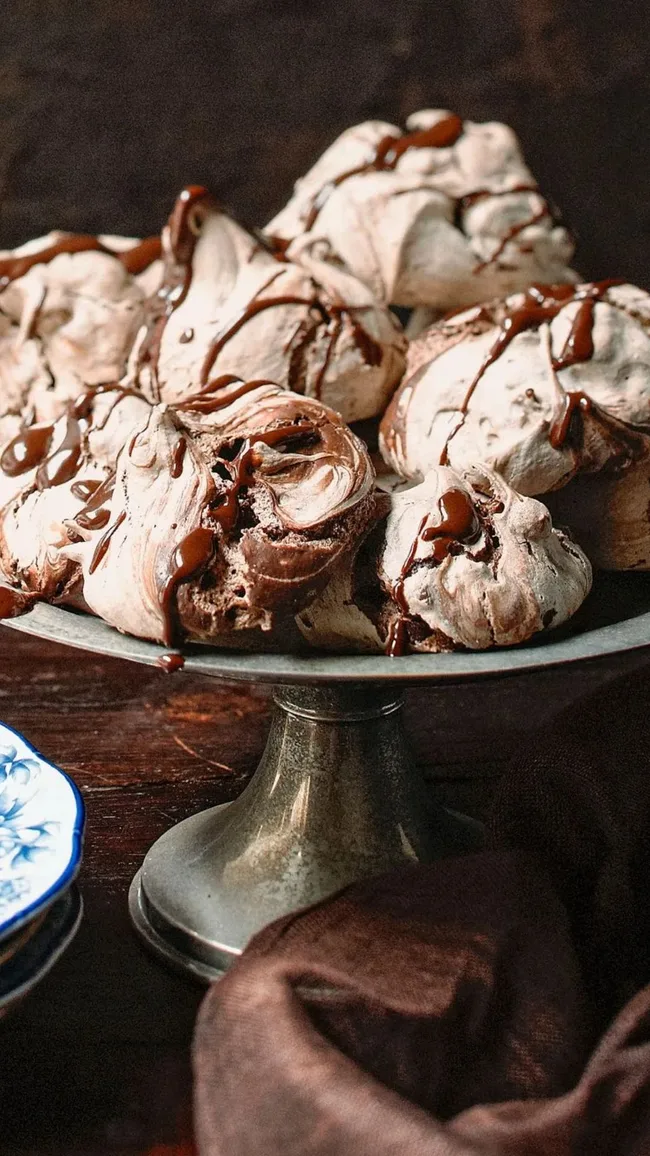 A dozen chocolate meringues on a vintage silver cake stand beside blue china plates on top of a dark timber tabletop