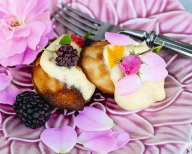 Honey madeleines with blackberries and cream on a purple flower plate topped with purple and pink flowers, sitting on a light blue linen tablecloth