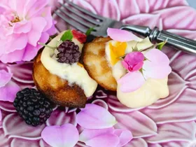 Honey madeleines with blackberries and cream on a purple flower plate topped with purple and pink flowers, sitting on a light blue linen tablecloth