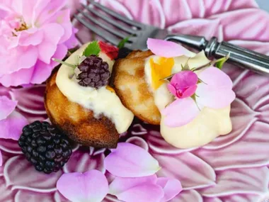 Honey madeleines with blackberries and cream on a purple flower plate topped with purple and pink flowers, sitting on a light blue linen tablecloth