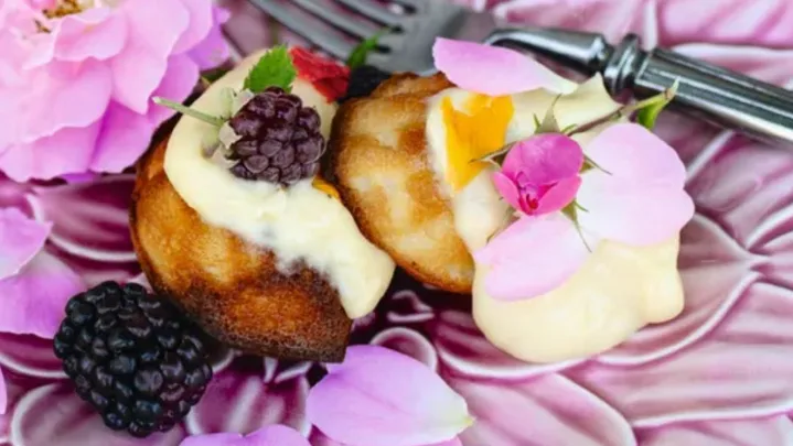 Honey madeleines with blackberries and cream on a purple flower plate topped with purple and pink flowers, sitting on a light blue linen tablecloth