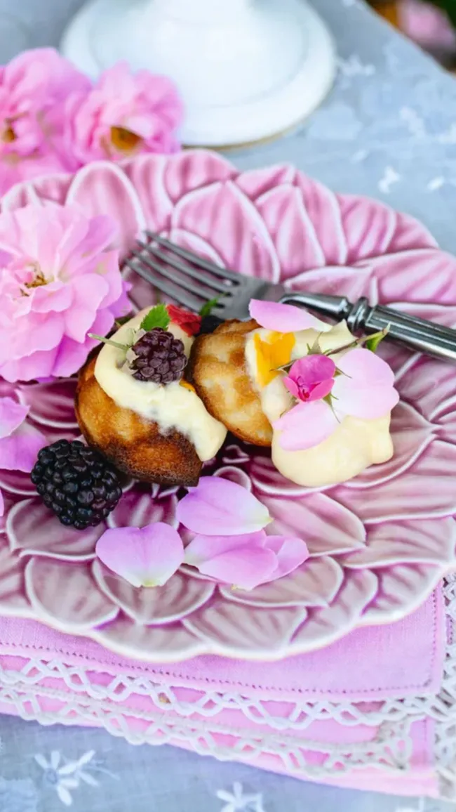 Honey madeleines with blackberries and cream on a purple flower plate topped with purple and pink flowers, sitting on a light blue linen tablecloth