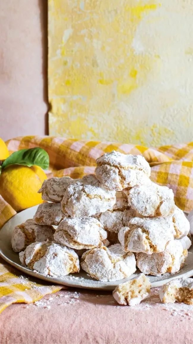 Lemon and fennel seed amaretti on a white plate on a yellow gingham table cloth with a bunch of lemons to the side