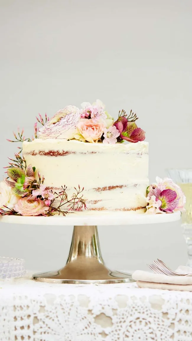 A passionfruit naked cake lightly frosted with white icing and decorated withpink and white florals on a cake stand