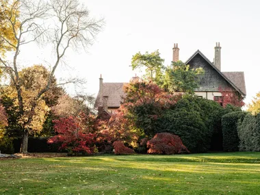 Wombat Park Estate in Daylesford. Arts and Crafts style grand building is just visible over hedges and bushes. From the left side of the photo, the trees and plantings are glorious shades of red, yellow and orange, with evergreen bushes still green on the right hand side.