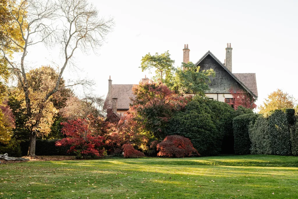 Wombat Park Estate in Daylesford. Arts and Crafts style grand building is just visible over hedges and bushes. From the left side of the photo, the trees and plantings are glorious shades of red, yellow and orange, with evergreen bushes still green on the right hand side.