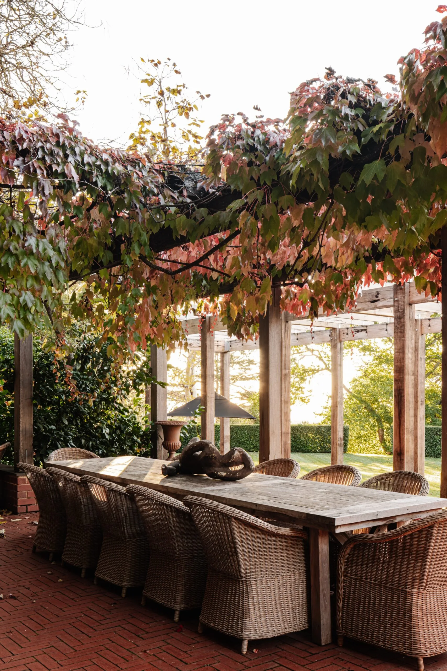 Outdoor dining area with wicker chairs, wooden table, and wooden arbour covered in autumn leaves, set in a lush garden setting.