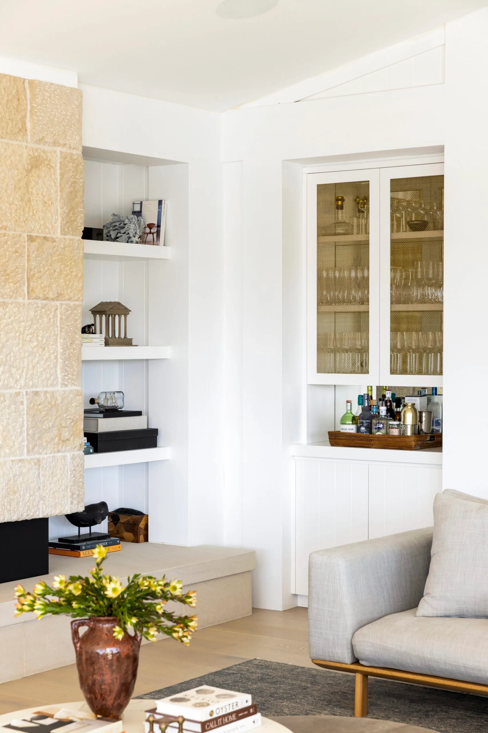 Living room in a fully redesigned and renovated heritage home with stone fireplace, white shelves, decorated cabinet, grey couch, and a vase of yellow flowers.