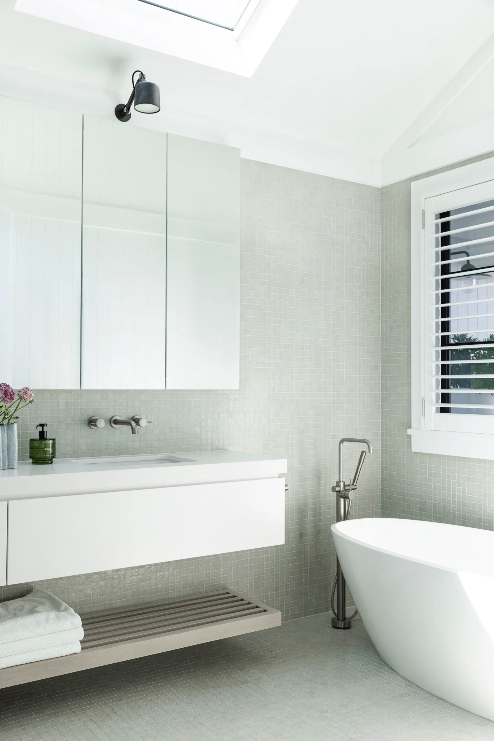 Bathroom with white vanity, freestanding tub, skylight, and wall-mounted faucet, featuring neutral tones.