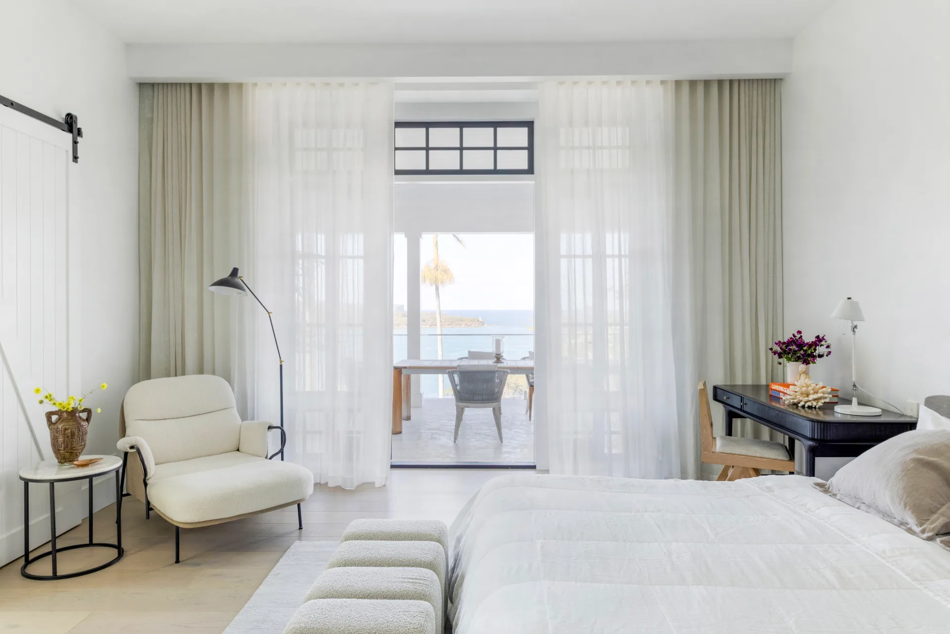 Cosy bedroom inside a fully renovated heritage home with white bedding, armchair, side table, and desk against sheer curtains revealing a balcony and ocean view.