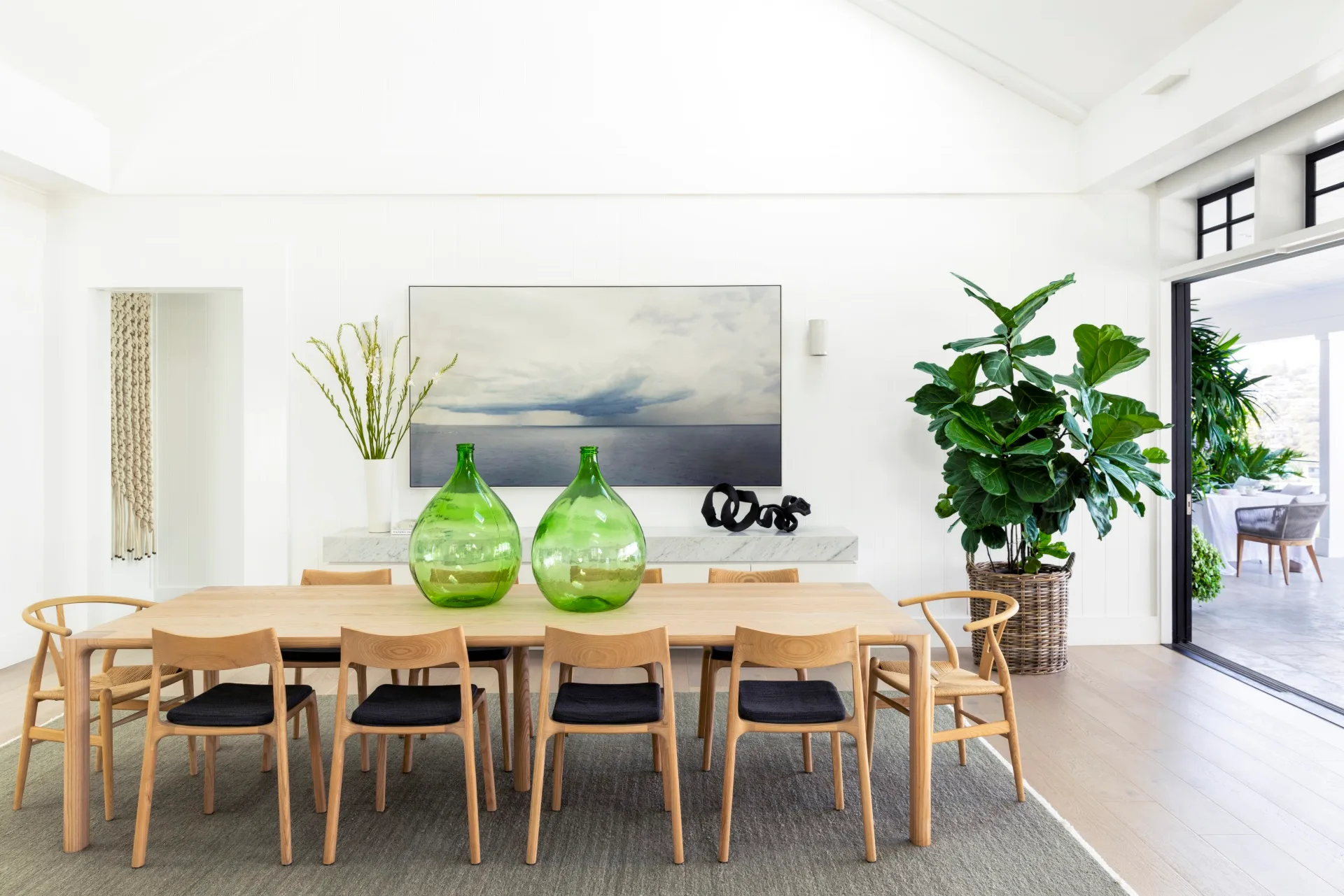 Bright dining room with wooden furniture, two large green glass vases on wooden dining table, a landscape painting, and lush indoor plants. A sliding door, to the right, opens out to a balcony.