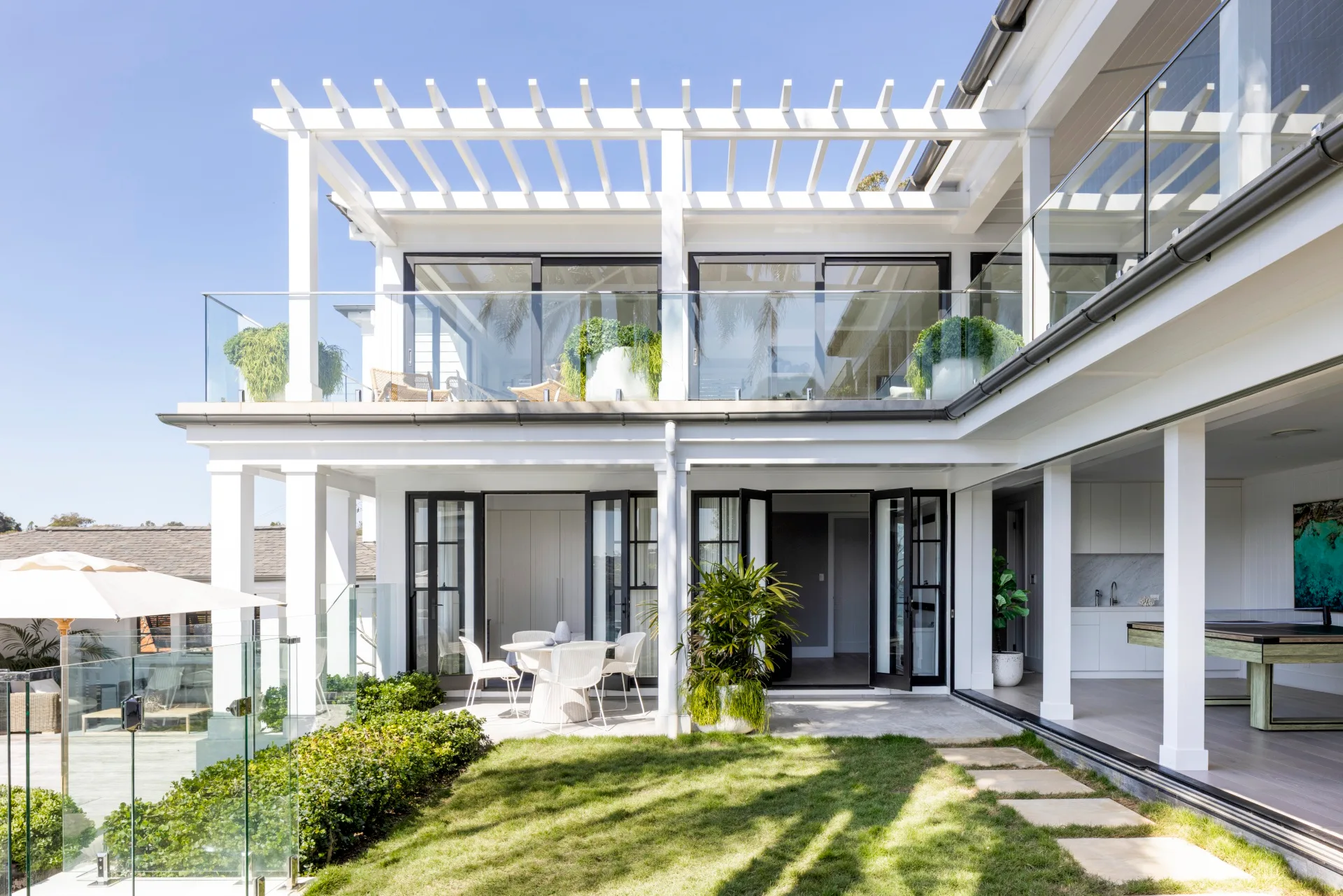 Exterior of a fully renovated and redesigned heritage home in a waterfront location. A two-story house with glass railings, white pergola, and garden patio under a clear blue sky.