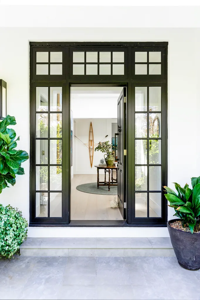 Open black-framed glass door with black-framed window panels on either side, potted plants on either side, leading to a bright hallway with a table and decor.