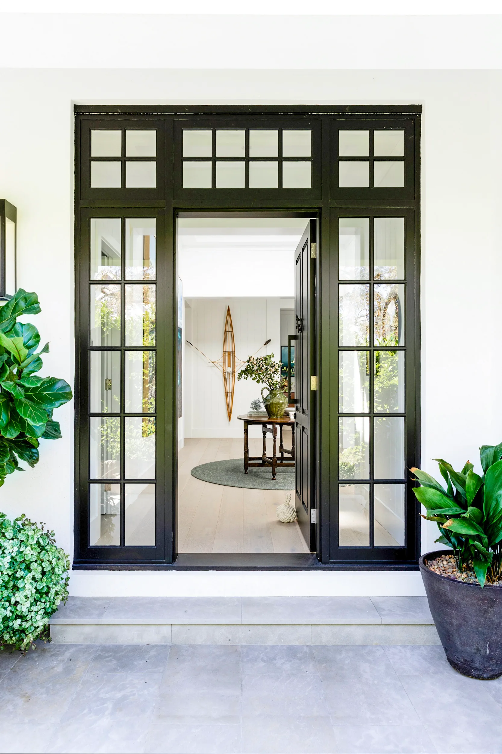 Open black-framed glass door with black-framed window panels on either side, potted plants on either side, leading to a bright hallway with a table and decor.