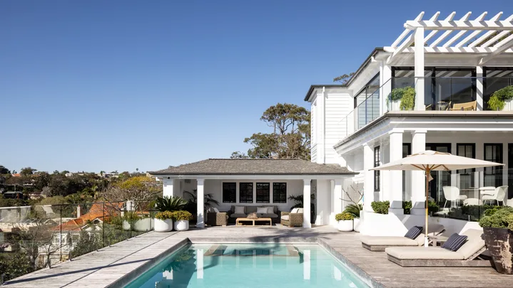 Luxurious house with a spacious patio, swimming pool, sun loungers, and pergola under a clear blue sky.