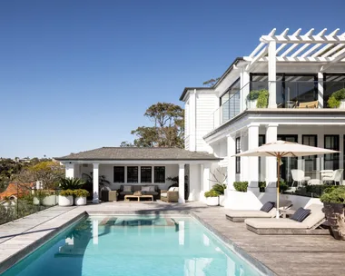 Luxurious house with a spacious patio, swimming pool, sun loungers, and pergola under a clear blue sky.