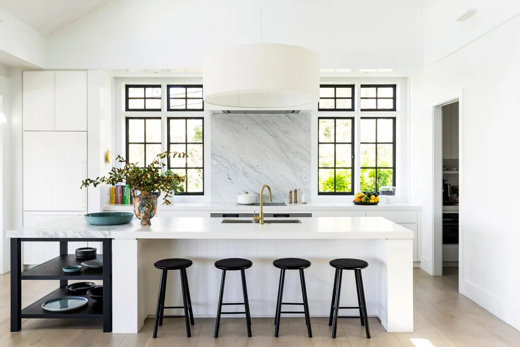 kitchen inside a fully redesigned and renovated heritage home with white island, four black stools, large central light fixture, and windows with black frames.