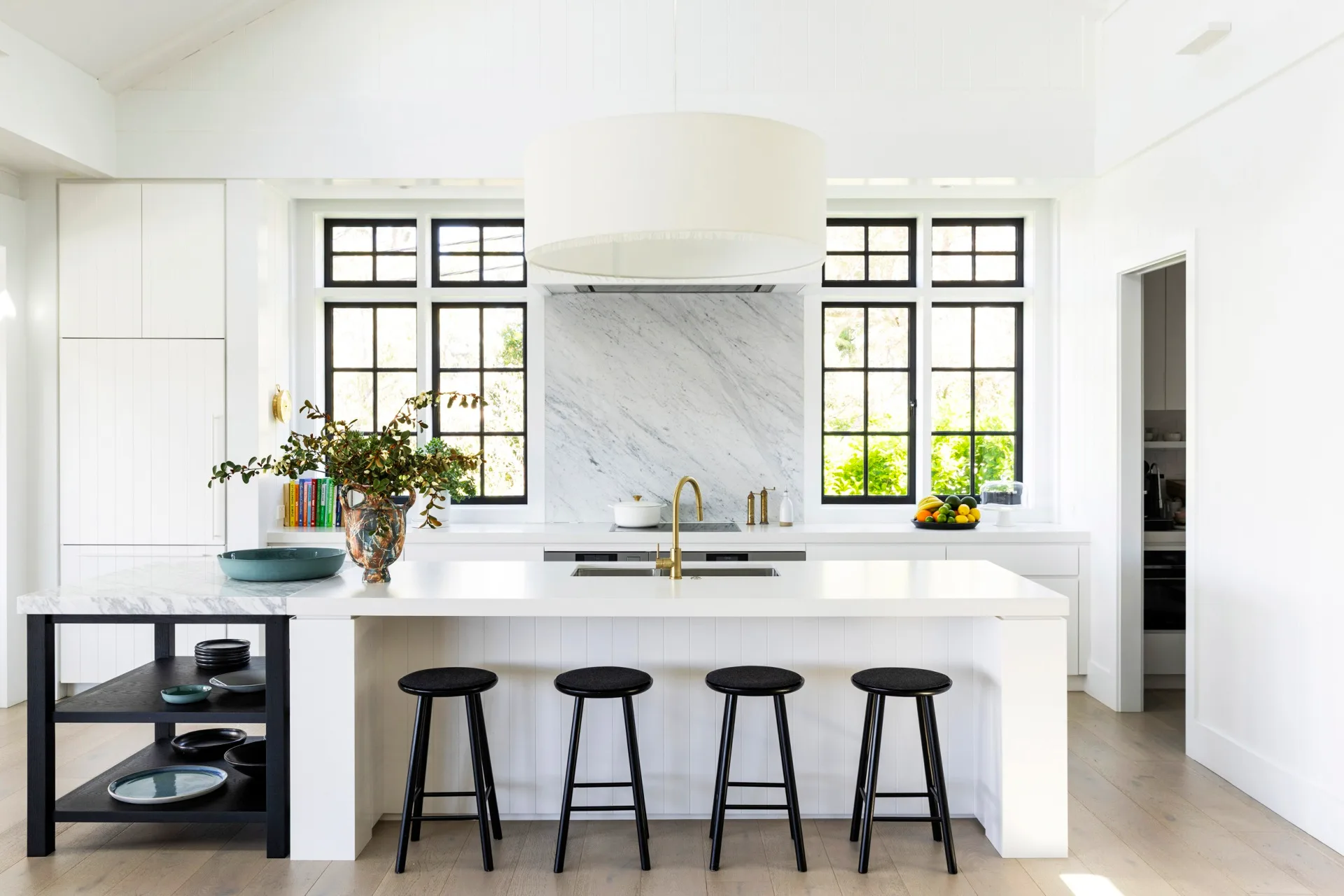 kitchen inside a fully redesigned and renovated heritage home with white island, four black stools, large central light fixture, and windows with black frames.