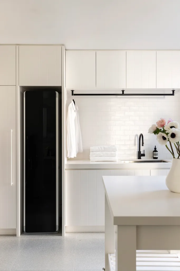 White laundry inside a fully renovated and redesigned heritage home. The laundry has stacked towels, a hanging shirt, black faucet, and flowers in a vase.