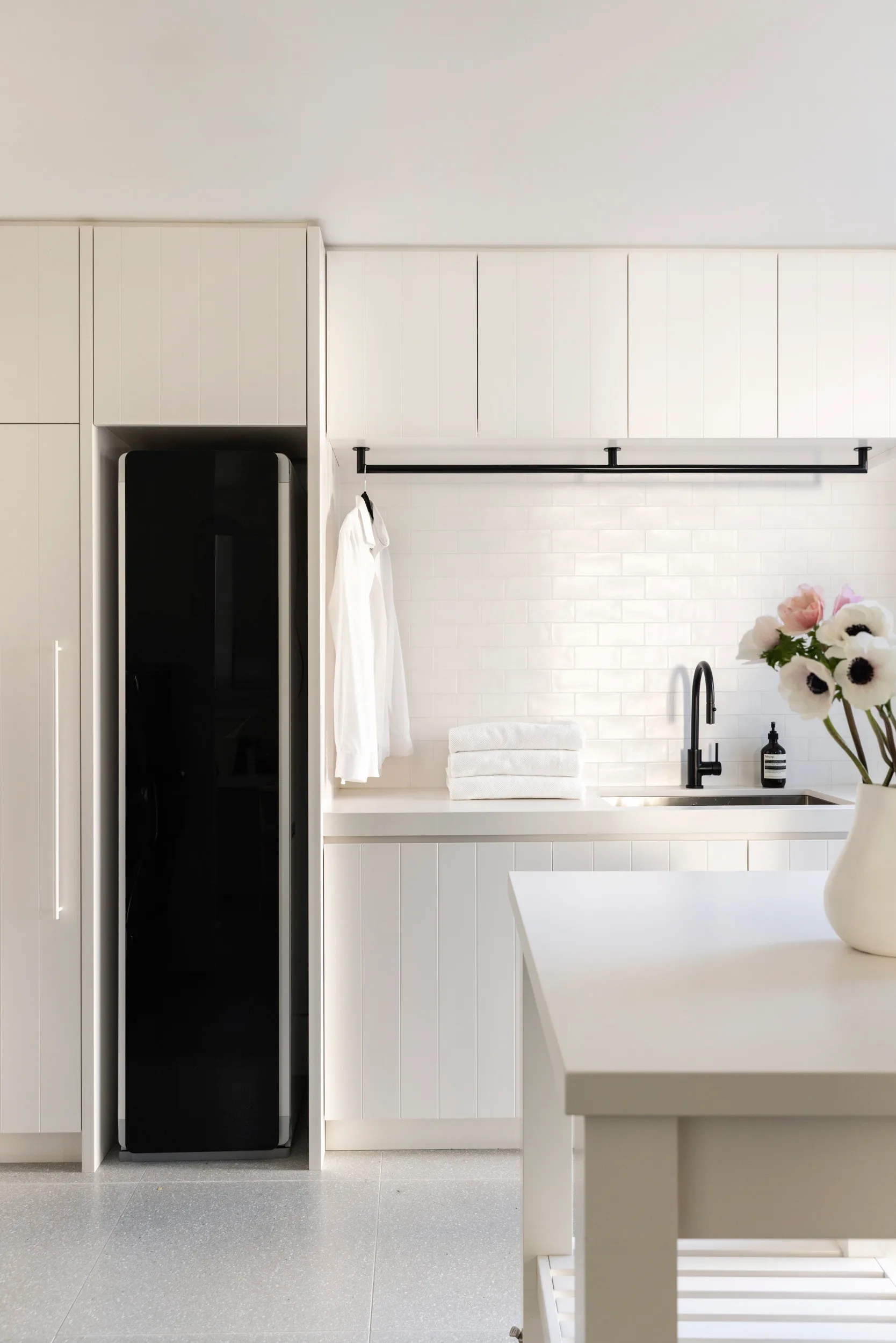 White laundry inside a fully renovated and redesigned heritage home. The laundry has stacked towels, a hanging shirt, black faucet, and flowers in a vase.