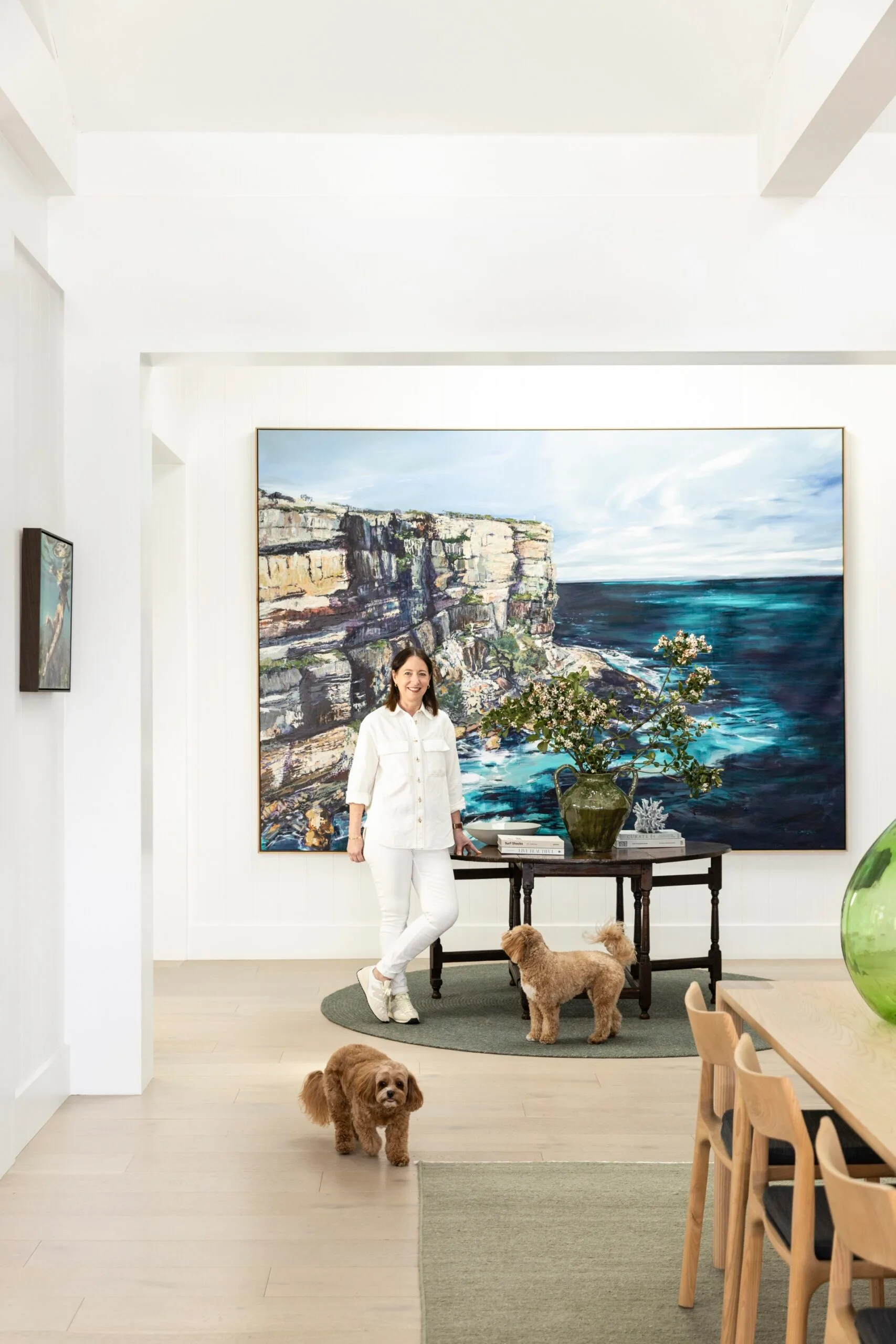 Person in white outfit with two fluffy brown dogs in a modern room, large seascape painting on the wall, light wood flooring.
