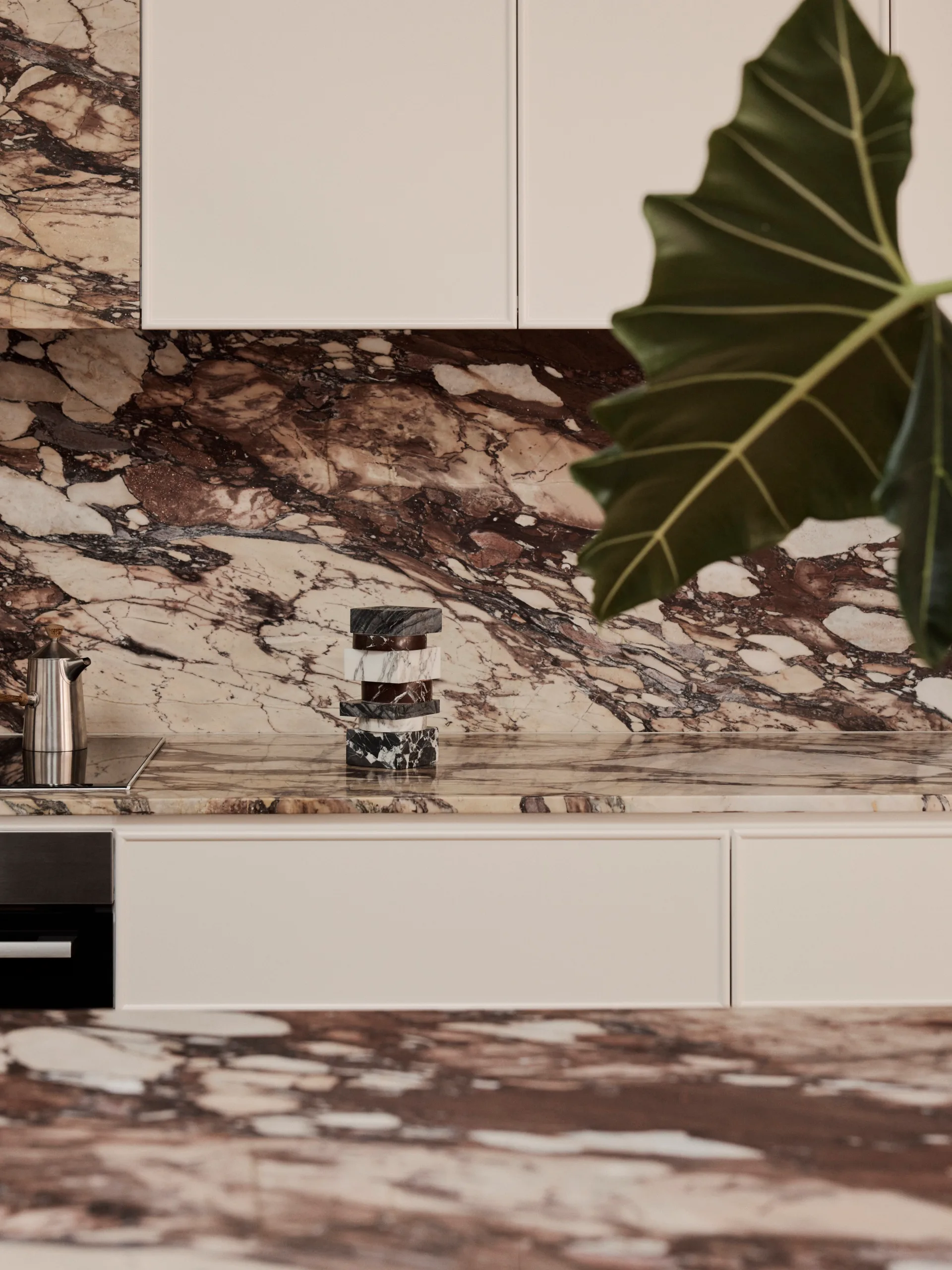 A close up photo of a galley kitchen. The top of the kitchen island is in the foreground and the material is marble with burgundy veining. This same marble has been used on the benchtop and splashback that are positioned across from the island. Joinery is white.