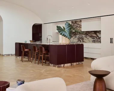 A contemporary galley kitchen in a Federation-era home with burgundy colour island profile, marble benchtop and splashback with burgundy veining and a white, arched high ceiling. Two sofas, facing each other, are just visible in the foreground as this is an open plan kitchen, dining and living space.
