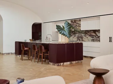 A contemporary galley kitchen in a Federation-era home with burgundy colour island profile, marble benchtop and splashback with burgundy veining and a white, arched high ceiling. Two sofas, facing each other, are just visible in the foreground as this is an open plan kitchen, dining and living space.
