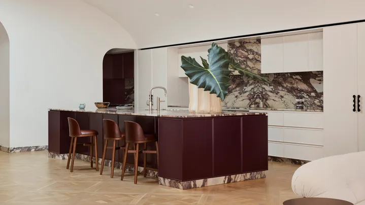 A contemporary galley kitchen in a Federation-era home with burgundy colour island profile, marble benchtop and splashback with burgundy veining and a white, arched high ceiling. Two sofas, facing each other, are just visible in the foreground as this is an open plan kitchen, dining and living space.