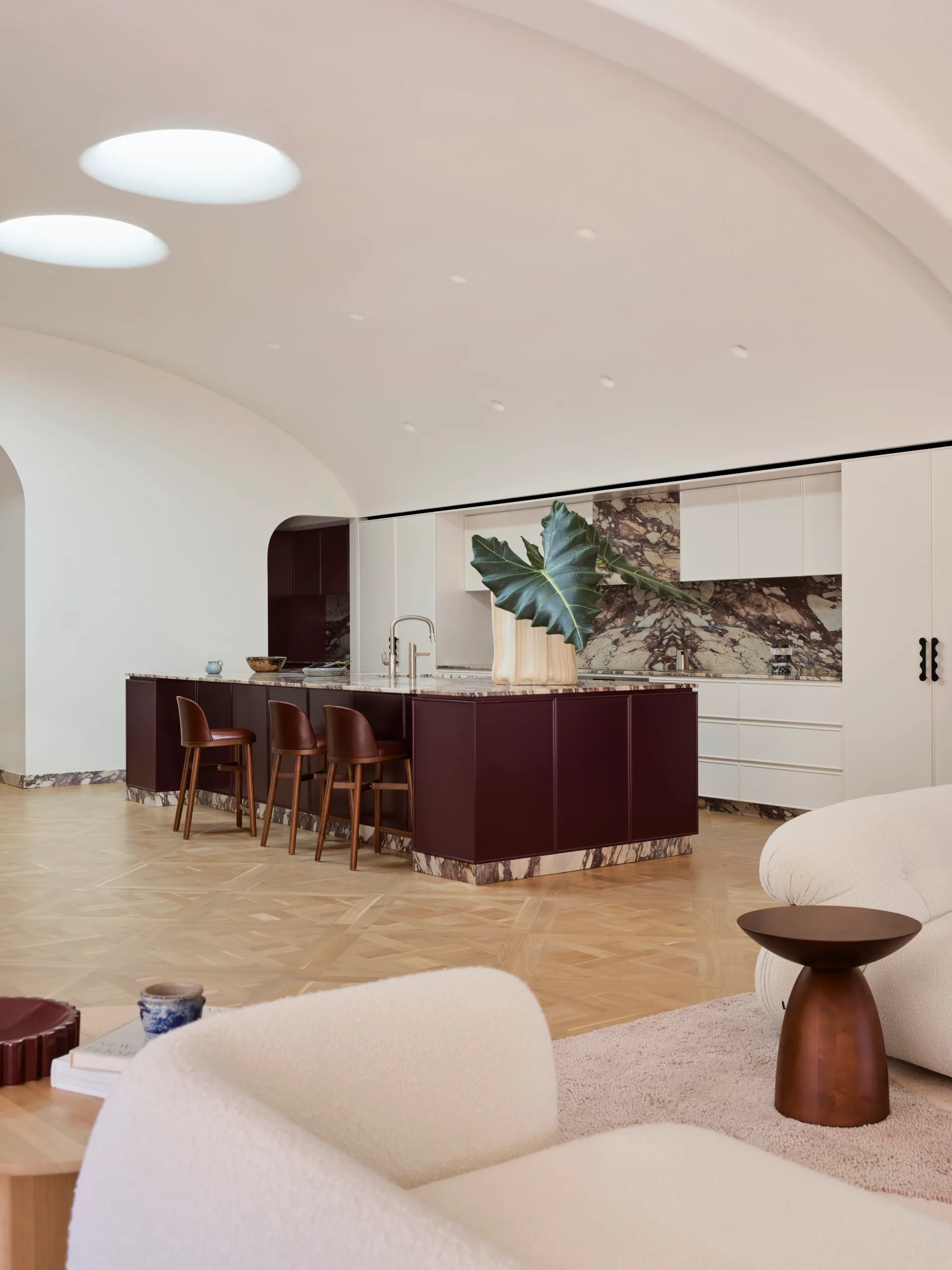 A contemporary galley kitchen in a Federation-era home with burgundy colour island profile, marble benchtop and splashback with burgundy veining and a white, arched high ceiling. Two sofas, facing each other, are just visible in the foreground as this is an open plan kitchen, dining and living space.