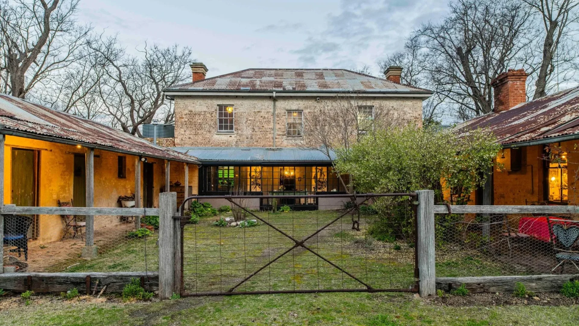 Historic Georgian two-story stone house with weathered metal roof, surrounded by trees and rustic wooden fencing.