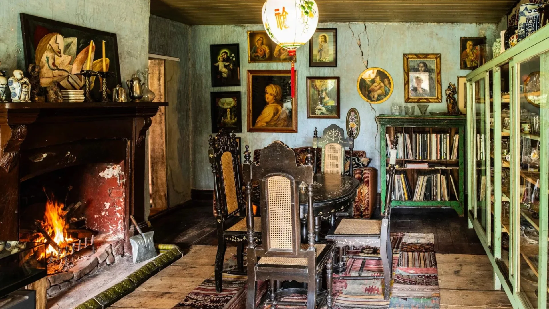 Cozy room inside a Georgian house with a fireplace, wooden table, chairs, and art-decorated walls. A bookshelf and green cabinet are visible.