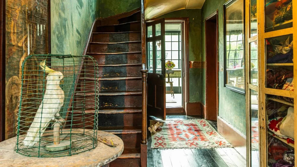 A taxidermied cockatoo in a cage on a table, near worn stairs and an open door in a vintage hallway with a rug.